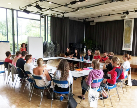 group of people seated around tables in a workshop presenter at a whiteboard by large windows in a bright room