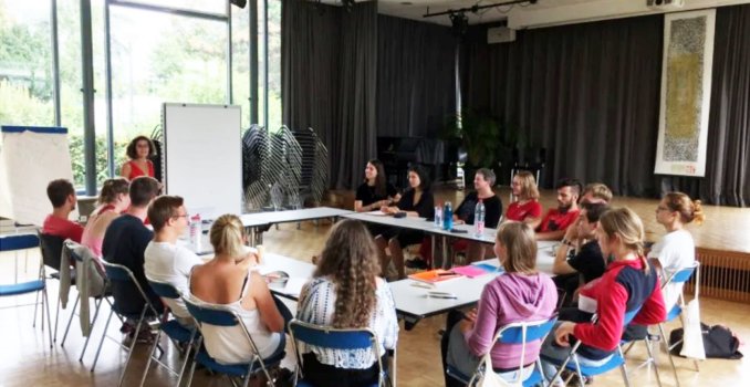 group of people seated around tables in a workshop presenter at a whiteboard by large windows in a bright room
