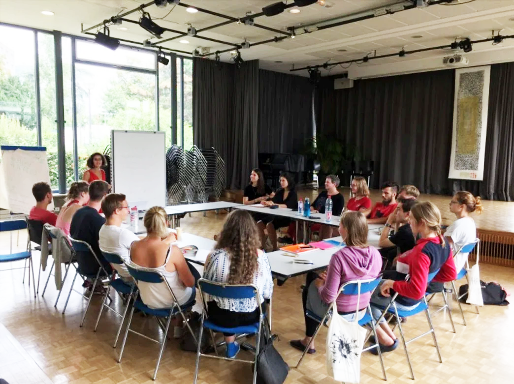 Group of people seated around tables in a workshop, presenter at a whiteboard by large windows in a bright room.