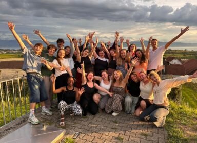 a large group of diverse friends on a terrace arms raised smiling for a team or group photo with hills and a lake in the background at sunset