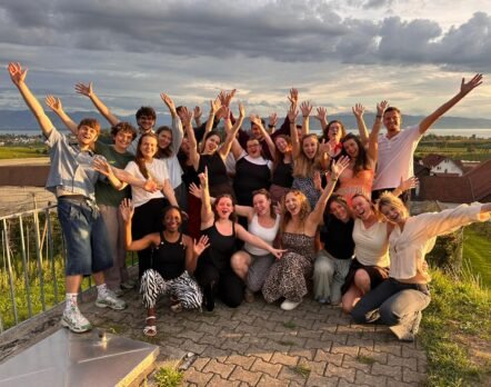 a large group of diverse friends on a terrace arms raised smiling for a team or group photo with hills and a lake in the background at sunset