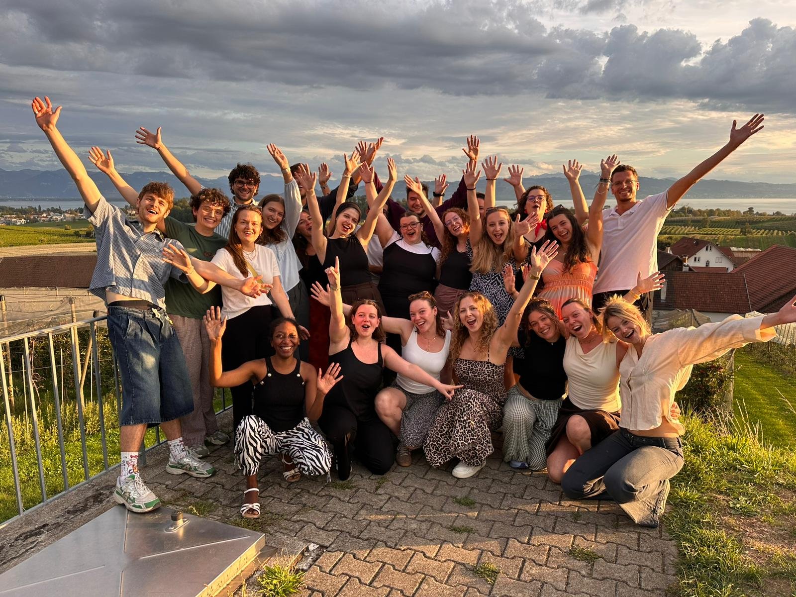 A large group of diverse friends on a terrace, arms raised, smiling for a team or group photo with hills and a lake in the background at sunset.